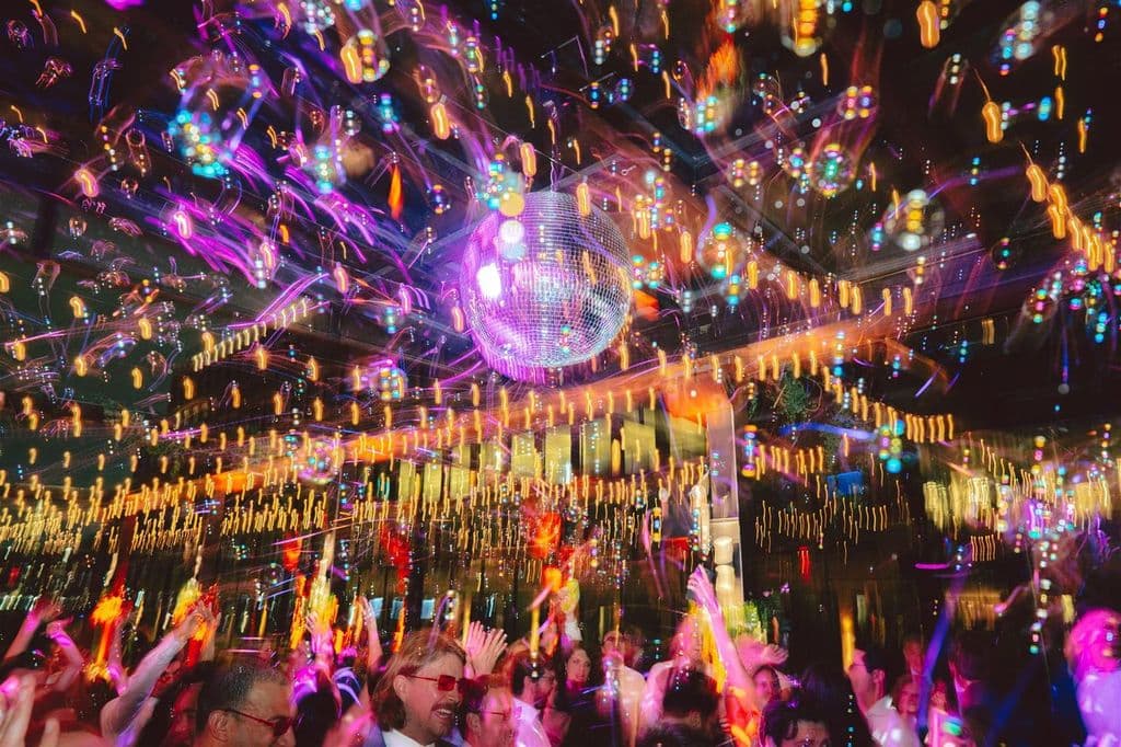 A packed dance floor lit by a mirror ball and streaks of coloured light