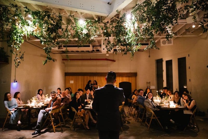 An MC addressing seated guests beneath a hanging greenery installation