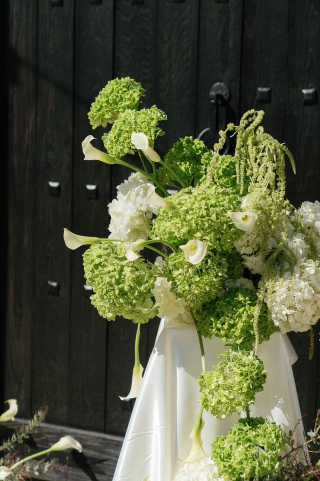A green-and-white arrangement of hydrangea and calla lilies on a draped pedestal