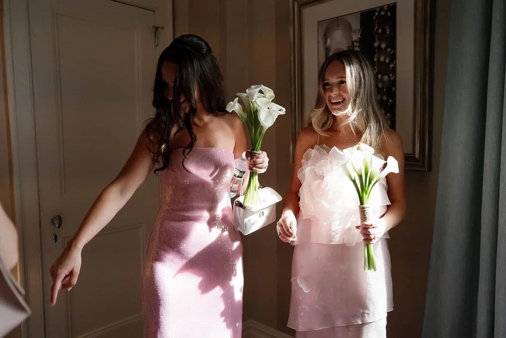Two guests in pink dresses laughing as they carry calla lily bouquets